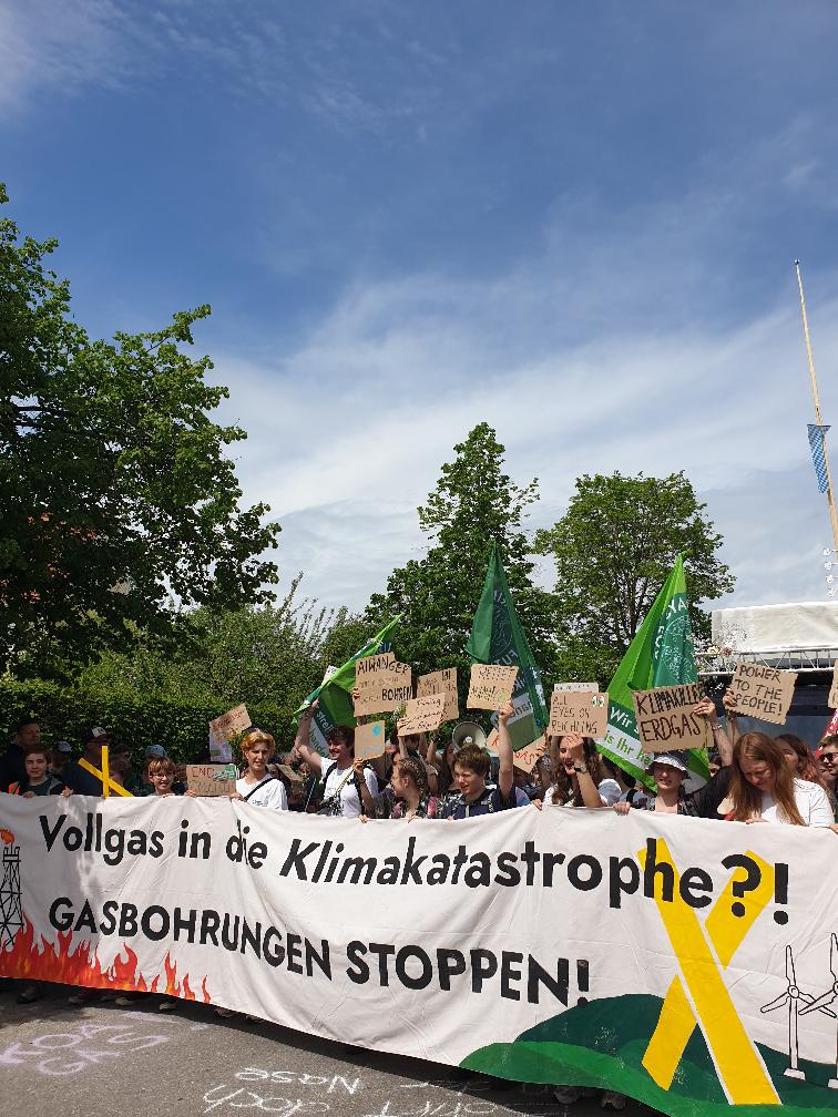 Ganz vornen in der Demo gab es ein straßenbreites Banner mit Zeichnungen von Feuer und Ölbohrtürmen auf der einen Seite und grüner Landschaft und Windrädern auf der anderen Seite. Dazu steht auf dem Banner die Aufschrift „Vollgas in die Klimakatastrophe?! GASBOHRUNGEN STOPPEN!“. Das Banner halten überwiegend Kinder und junge Menschen. Dahinter werden viele weitere FFF-Flaggen und Pappschilder in die Höhe gehalten mit Sprüchen wie „KLIMAKILLER ERDGAS“ und „POWER TO THE PEOPLE“. Im Hintergrund sind Bäume und der Maibaum von Reichling.