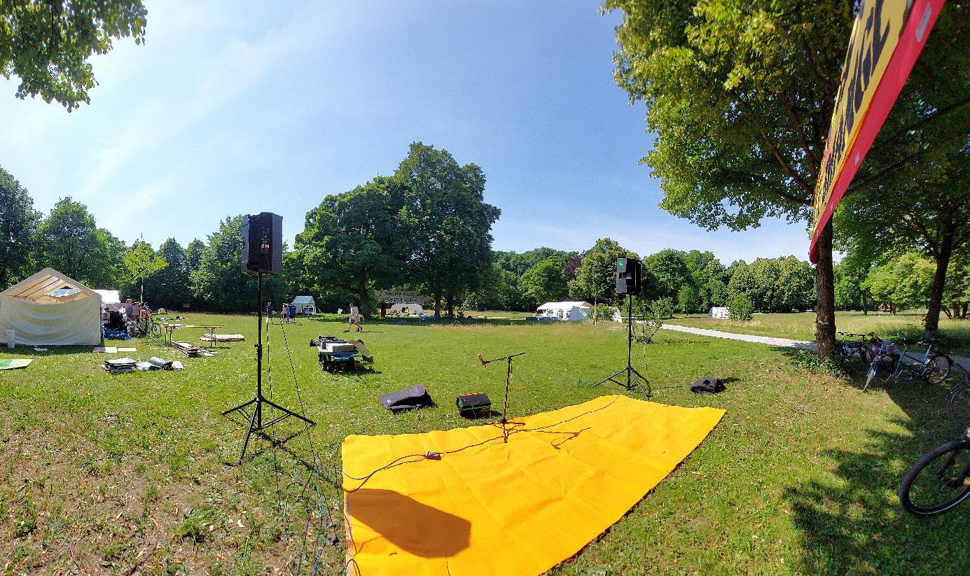 Eine große Wiese mit großen Bäumen. Dazwischen werden Zelte aufgebaut. Im Vordergrund ist Tontechnik, wie große Lautsprecherboxen.