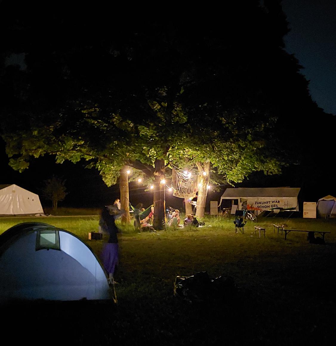Das Bild zeigt eine Gruppe von großen Laubbäumen in der dunklen Nacht. Zwischen den Bäumen sitzen junge Menschen, beleuchtet durch einen Kranz von Lampen, der zwischen den Bäumen hängt. Im Umfeld stehen Zelte und Bierbänke.