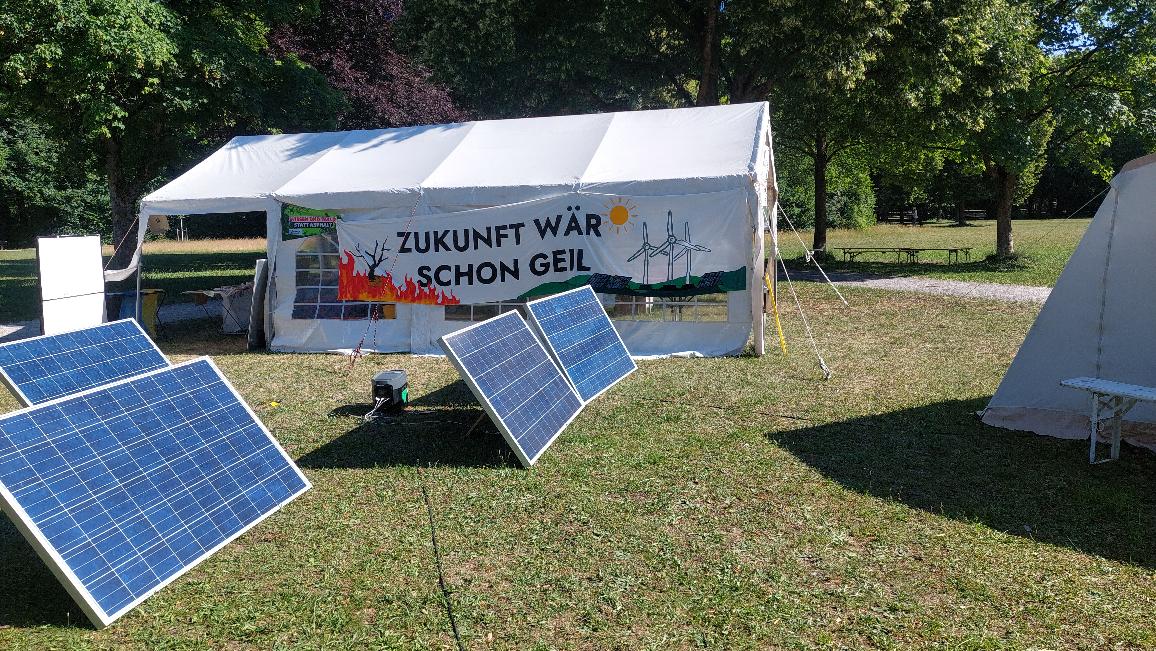 Das Bild zeigt vier Solarplatten sowie ein paar Zelte auf einer sonnenbeschienen Wiese und große Laubbäume im Hintergrund.